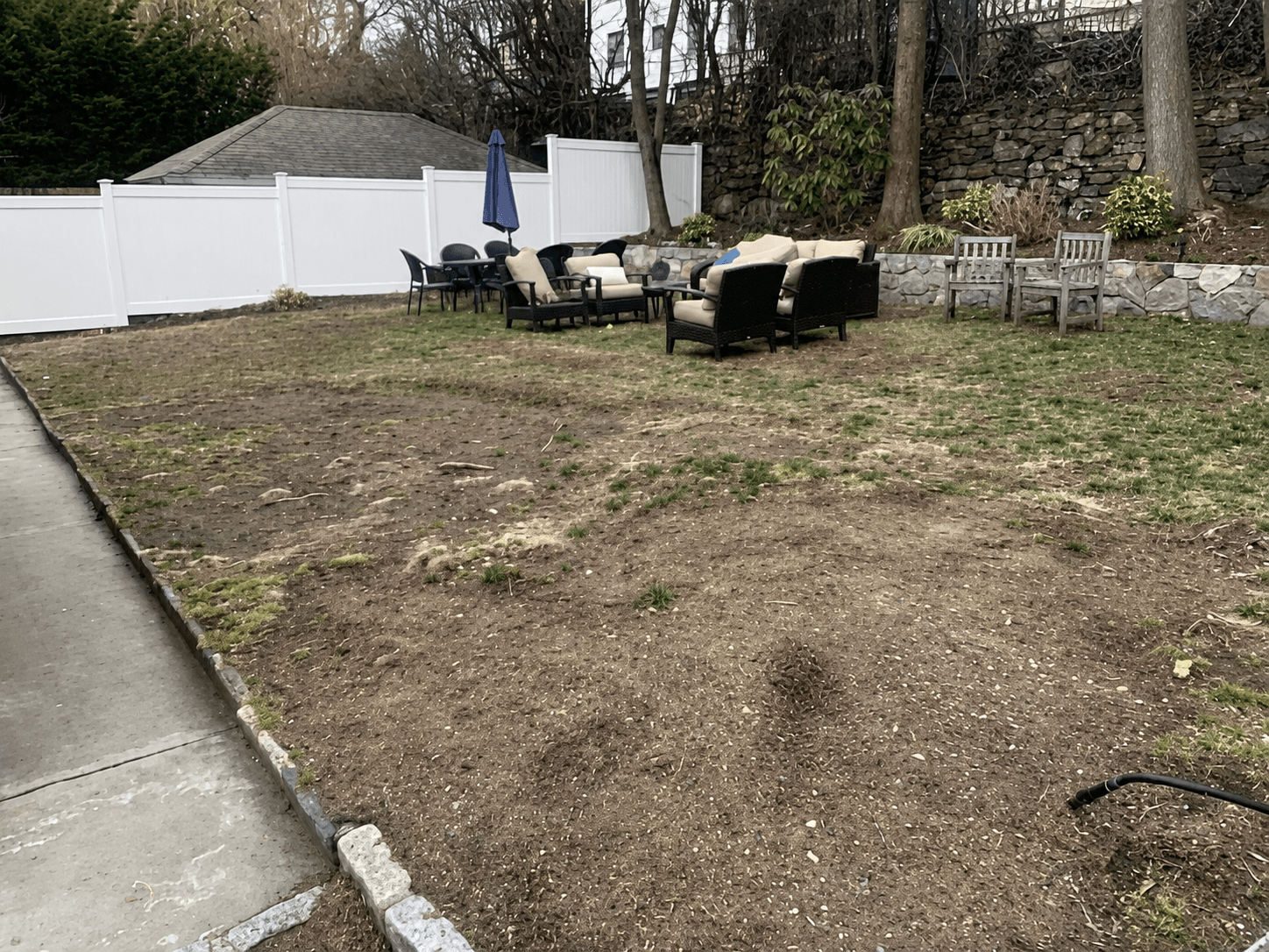 Patchy dirt-and-grass backyard before the renovation, with scattered patio furniture against a white vinyl fence and stone retaining wall